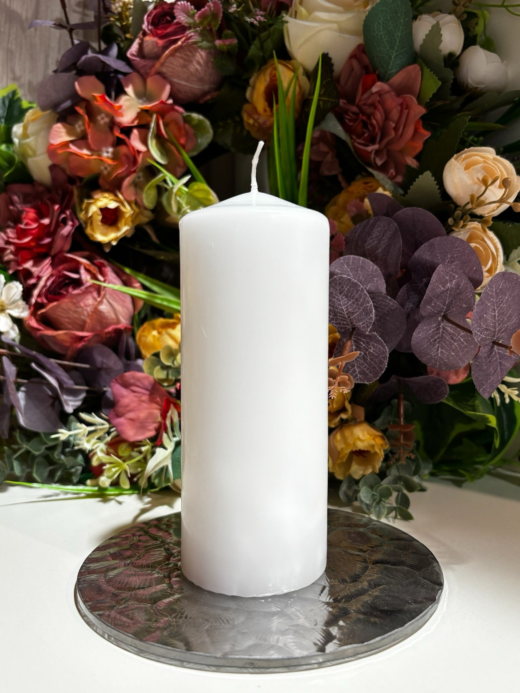 Candles Plus white pillar candle on a textured silver base, set against a backdrop of colourful artificial flowers.